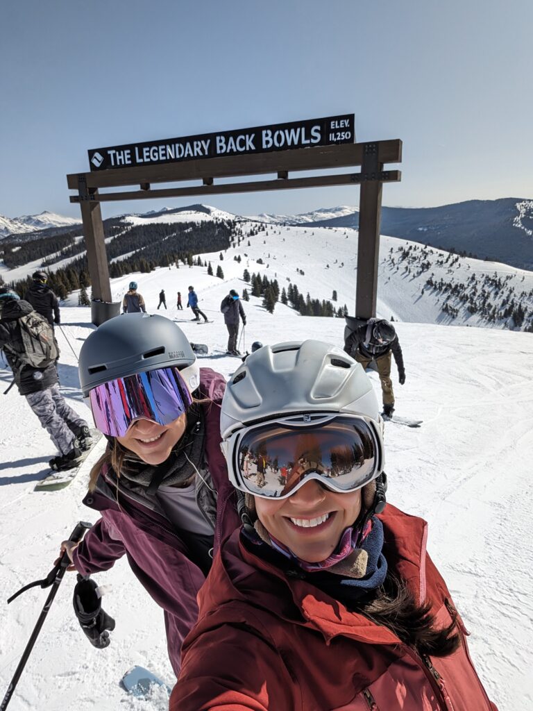2 women smiling at the top of a mountain while skiing